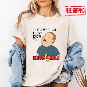 May include: A cream-coloured t-shirt featuring a cartoon image of Bobby Hill from King of the Hill. The shirt has the quote "THAT'S MY PURSE! I DON'T KNOW YOU!" printed above the character. The King of the Hill logo is below the character.