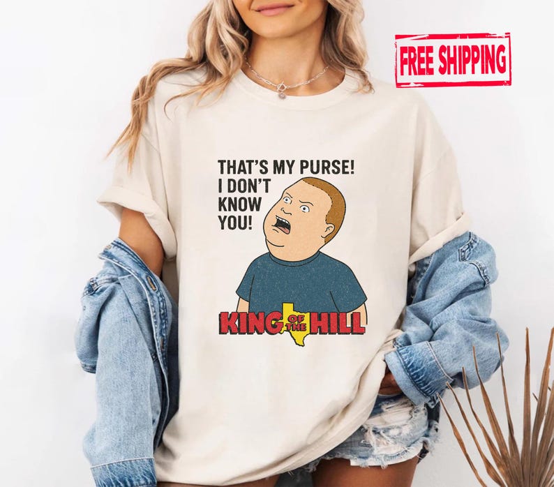 May include: A cream-coloured t-shirt featuring a cartoon image of Bobby Hill from King of the Hill. The shirt has the quote "THAT'S MY PURSE! I DON'T KNOW YOU!" printed above the character. The King of the Hill logo is below the character.
