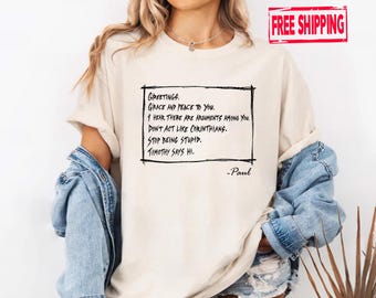 May include: Cream-colored t-shirt with a black handwritten text box containing a quote. The text reads "Greetings, Grace and Peace to You. I hear there are arguments among you. Don't act like Corinthians. Stop being stupid. Timothy says hi."