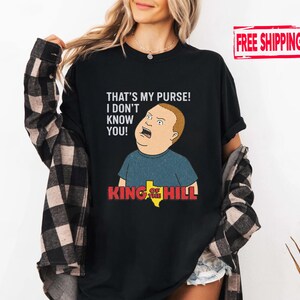 May include: Black t-shirt with a cartoon image of Bobby Hill from King of the Hill. The shirt has the text "THAT'S MY PURSE! I DON'T KNOW YOU!" above the character and "KING OF THE HILL" below.