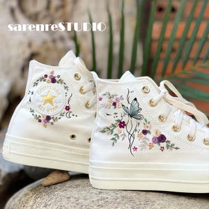 May include: Cream-colored high-top sneakers with floral and butterfly embroidery. The sneakers feature a Converse logo with a gold star and are adorned with purple, pink, and green floral designs. The text "sarenreSTUDIO" is visible.