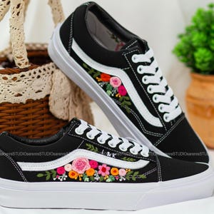 May include: Black canvas trainers with white soles and laces. The trainers have a white side stripe and are decorated with colourful floral embroidery in pink, orange, and green. A fashionable and unique footwear choice.