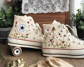 Puede incluir: Un par de zapatillas altas color crema con diseños florales e insectos bordados. Las zapatillas tienen cordones blancos, suela negra y una raya roja. El logo de Converse es visible.