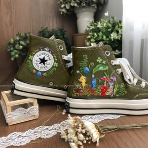 May include: Olive green high-top trainers with embroidered floral and mushroom designs. The left trainer features a Converse All Star logo. The right trainer has a colourful embroidered scene with cartoon characters and plants. White laces and soles.