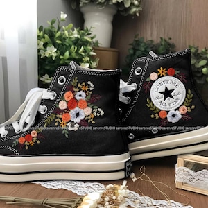 May include: Black high-top sneakers with floral embroidery. The sneakers feature white laces, a white rubber sole, and a classic Converse All Star logo patch. The embroidered flowers include orange, red, and white blooms.