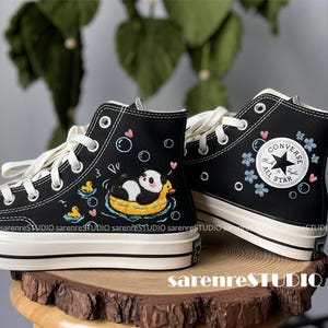 May include: Black high-top sneakers with white laces and soles. One shoe features a panda in a duck floatie, bubbles, and hearts. The other shoe has a Converse All Star logo with blue flowers. The brand name "sarenreSTUDIO" is printed on the side.