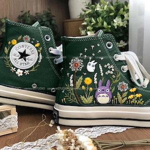 May include: Dark green high-top trainers with white laces and soles. The trainers feature embroidered floral and cartoon character designs. One trainer has a Converse All Star patch surrounded by flowers. The other has a Totoro design.