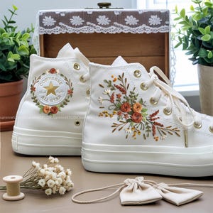Peut inclure: Baskets montantes blanches avec broderies florales. L'une arbore le logo Converse All Star avec une couronne florale. L'autre présente un motif floral avec des roses orange et rouges. Lacets blancs, œillets dorés.