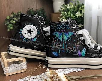 May include: Black high-top sneakers with white laces and soles. One shoe features the Converse All Star logo, the other a colorful moth and crystal design. The shoes are decorated with celestial and botanical elements.