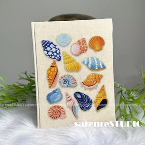 Puede incluir: Un diario cubierto de tela beige con diseños de conchas bordadas en varios colores. Las conchas incluyen azul, naranja, amarillo y rosa, creando un tema costero. El texto "sarenreSTUDIO" está impreso en la parte inferior.