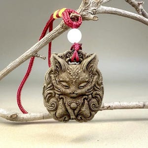 May include: A detailed, tan-colored pendant featuring a stylized fox face with intricate carvings. The pendant hangs from a red cord with a white bead and yellow accents. The fox's features include closed eyes and detailed fur patterns.