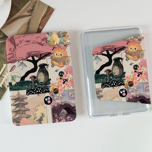 May include: Two phone cases with a colourful Japanese anime design. The design features characters, cherry blossoms, and traditional Japanese architecture. One case is inside a clear protective case.