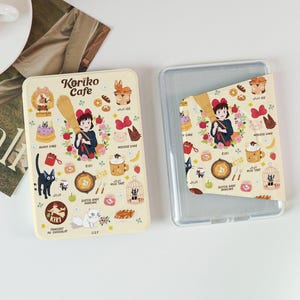 May include: Two phone cases with a cream-colored design featuring the text "Koriko Cafe" and illustrations of a girl, a black cat, cakes, and other food items. One case is open, revealing the interior.