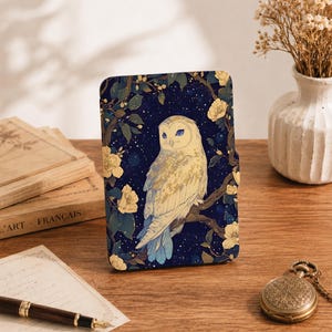 Op de afbeelding: Een rechthoekige e-readerhoes met een witte uil op een tak tegen een donkerblauwe sterrenhemel. Het ontwerp bevat gele bloemen en groene bladeren, met goudkleurige accenten op de veren van de uil.
