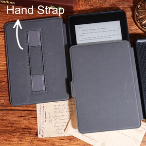 May include: A gray e-reader case with a hand strap, open to display the device. The case is made of a smooth material and has a simple design. The e-reader displays text. The words "Hand Strap" are visible.