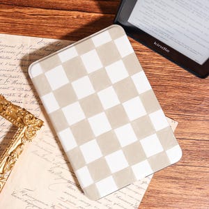May include: A rectangular Kindle case with a beige and white checkered pattern. The case is lying on top of a vintage document with a gold frame in the background. A Kindle e-reader is visible in the background.