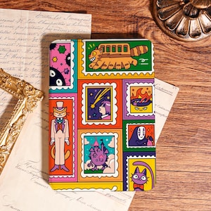 May include: A colorful notebook featuring a collage of cartoon illustrations in a postage stamp design. The images include a cat in a suit, a bus-like creature, a girl watching a shooting star, and a purple cat with a bow tie. The notebook is on a wooden surface.