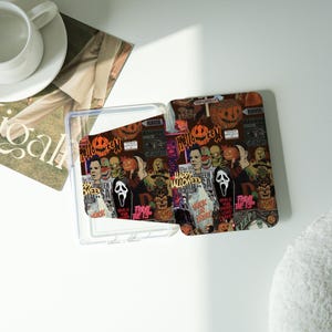 Puede incluir: Una funda de teléfono rectangular transparente con un diseño de Halloween. El diseño presenta imágenes de calabazas, calaveras y personajes de películas de terror. La funda está abierta.