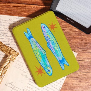 May include: A Kindle cover with a vibrant yellow-green background featuring two stylized fish in shades of blue and green, accented with orange starbursts. The cover is rectangular and rests on a wooden surface.