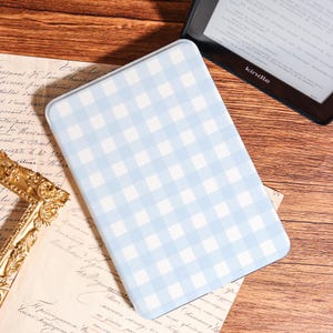 Peut inclure: Un étui rectangulaire à carreaux vichy bleu clair et blanc, probablement pour une liseuse ou une tablette, repose sur une surface en bois. Une liseuse Kindle est visible en arrière-plan. L'étui est de couleur pastel.