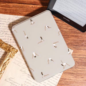 May include: A light beige Kindle cover featuring a pattern of yoga poses in shades of brown and white. The cover is rectangular and appears to be made of a smooth material. The Kindle is black.