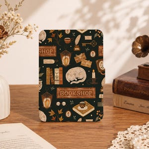 May include: A rectangular e-reader case featuring a dark green background with a repeating pattern of sleeping cats, lanterns, books, envelopes, keys, and a sign that reads BOOKSHOP. The design includes warm brown, cream, and gold-toned illustrations.