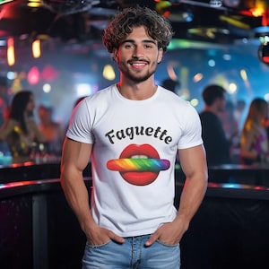 May include: White t-shirt with the word "Faguette" in black script above a graphic of red lips and a rainbow-coloured baguette. The person is wearing blue jeans and is smiling. The background is a blurred nightclub scene.
