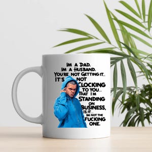 May include: White ceramic mug with a black and blue graphic. The mug features a man in a blue hoodie and text that reads "IM A DAD. IM A HUSBAND. YOU'RE NOT GETTING IT.. IT'S NOT CLOCKING TO YOU... THAT I'M STANDING ON BUSINESS, IS IT... IM NOT THE FUCKING ONE."