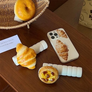 May include: A white iPhone case with a croissant design. The case has a white background with a brown croissant on the front. The text "is killing" is printed on the case. The case is on a wooden table with a white phone stand and a pastry.