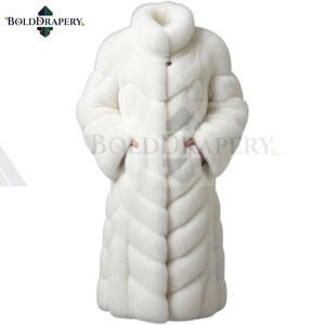 May include: A full-length, white fur coat with a high collar and a button closure. The coat features a chevron pattern and long sleeves. The brand name "BOLDDRAPERY" is visible in the upper left corner.