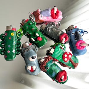 Pode incluir: Uma coleção de oito capas personalizadas para isqueiros feitas de argila polimérica, com vários designs esculpidos, incluindo monstros, rostos com lábios vermelhos, um cacto verde com flores coloridas e um cogumelo verde, cada um com isqueiro.