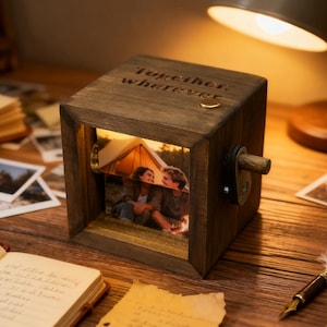 May include: A wooden photo cube with a picture of a couple inside, illuminated by a warm light. The cube has a crank on the side and the words "Together, wherever" engraved on top. The cube sits on a wooden surface with other photos and a lamp.
