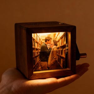 May include: A small, wooden box with a window showing a couple kissing in a library setting. The box has a crank and is held in a hand. The interior scene is lit with a warm glow, creating a romantic ambiance.