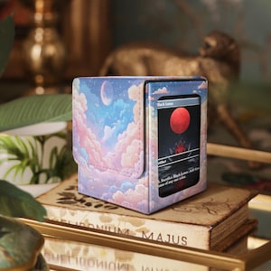 May include: A card storage box with a pastel sky design featuring clouds, a crescent moon, and birds. The box displays a card with a red orb and the text "Black Lotus". The box is on a stack of books.