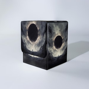 May include: A black card storage box with a dramatic, fantasy-themed design. The artwork features a dark, ominous landscape with a large, dark circular shape in the sky, and silhouetted figures. The box has a magnetic closure.