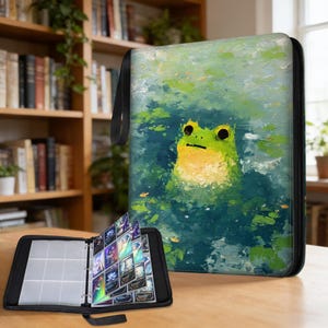 Könnte beinhalten: Schwarzer Kartenordner mit einem Froschgemälde auf dem Einband. Der Frosch ist grün und gelb, vor einem blau-grünen Hintergrund. Der Ordner ist geöffnet und zeigt transparente Kartenhüllen mit bunten Karten.