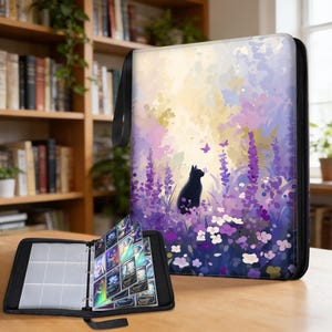 May include: Black card binder with a colorful floral design featuring a black cat, purple flowers, and butterflies. The binder is open, revealing clear card sleeves filled with trading cards. A black handle is attached to the binder.