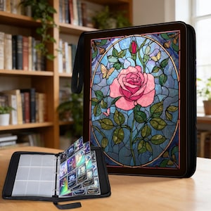 Puede incluir: Carpeta de tarjetas negra con un diseño de vidriera que presenta una rosa rosa, mariposas y hojas verdes. La carpeta está abierta, revelando fundas transparentes para tarjetas llenas de tarjetas. Un asa negra está unida al lateral.