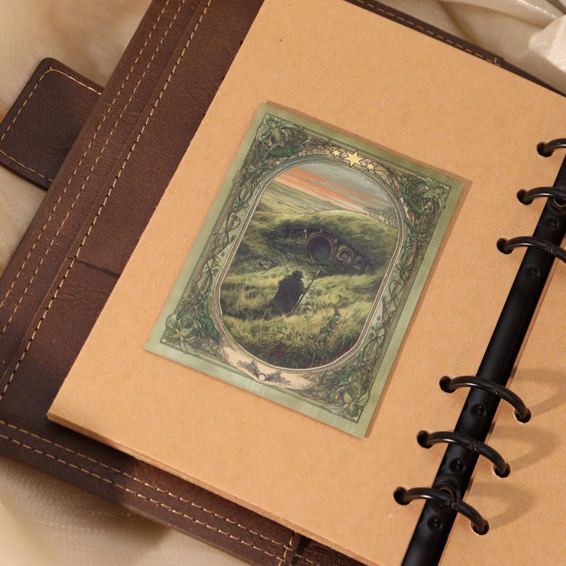May include: A rectangular sticker featuring an illustration of a figure walking toward a round-doored hillside dwelling. The scene is framed by an ornate green border with three stars at the top, placed on a tan page within a dark brown ring binder.