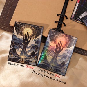 May include: Two rectangular prints with black frames featuring a surreal illustration of a giant hand reaching toward a glowing sun above a landscape. One print has a glossy finish, while the other features a holographic rainbow foil sheen.