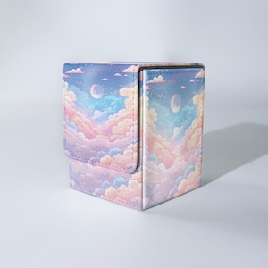 Puede incluir: Caja de almacenamiento cuadrada con cierre de solapa, decorada con una escena de cielo pastel. El diseño presenta nubes, una luna creciente y estrellas en tonos de azul, rosa y morado. Ideal para guardar objetos pequeños.