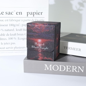 May include: A black and red card storage box with the text "Berserk Black Knight". The box features a sunset design with a dark sky and red water. The box is sitting on a grey book.