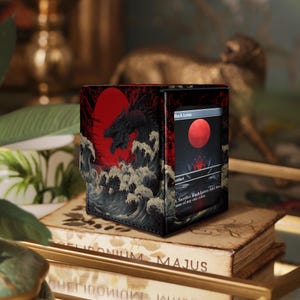 Könnte beinhalten: Schwarze und rote Kartenbox mit einem Drachendesign vor roter Sonne und Wellen. Die Box zeigt eine Karte mit rotem Orb und dem Text "Black Lotus". Die Box steht auf einem Bücherstapel.