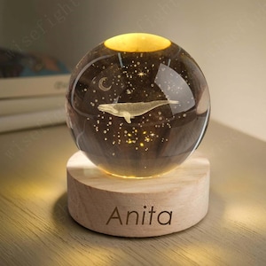 以下が含まれることがあります： クジラのデザイン、星、三日月が描かれた装飾的なガラス球。温かい光で照らされています。「Anita」という名前が刻まれた木製の台座の上にあります。