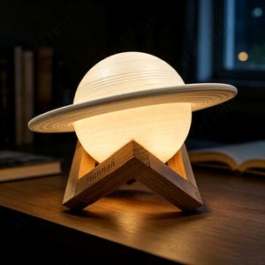 以下が含まれることがあります： 惑星の形をした球体のランプで、目立つリング構造を持ち、温かく柔らかな光を放っています。ランプはV字型のベースを持つ明るい色の木製スタンドの上に置かれており、木製の脚の一つにはHannahという名前が刻印されています。