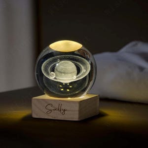 以下が含まれることがあります： 木製の台座の上に置かれた、惑星のデザインが施された透明なガラス球。惑星にはリングがあり、小さな球体で囲まれています。ベースには「Sally」という名前が刻まれています。球体は内側から照らされ、温かい光を放っています。