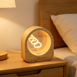 May include: A light-colored wooden lamp with a rounded arch shape and a circular cutout. Inside the cutout, a warm glowing neon-style light filament forms a heart shape. The name Oliver is engraved in dark letters on the front base of the lamp.
