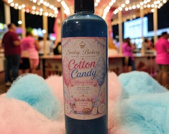 May include: A cylindrical bottle of blue liquid soap with a label reading Sudsy Bakery Cotton Candy Whimsy Wash, featuring illustrations of cotton candy and a carnival cart, with a volume of 16 fl oz (473 mL) printed on the front label.