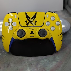 May include: A custom yellow PlayStation 5 controller with a black Wolverine logo. The controller has blue accents and black buttons. The design features the PlayStation logo and directional buttons.