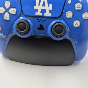 May include: A blue and black PlayStation 5 controller featuring the Los Angeles Dodgers logo. The controller has white buttons and a black grip. The LA logo is in white.